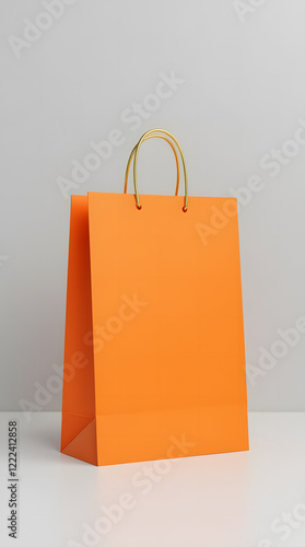 Bright orange shopping bag with gold handles on light grey background is a versatile template for showcasing branding or product design. Stylish and minimalistic look.