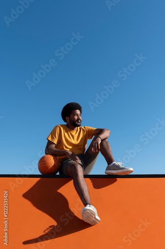 Afro man with basket ball sitting on orange wall under blue sky photo