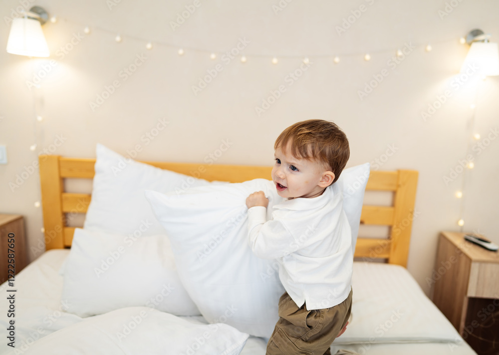 Young child plays joyfully with pillows. A cheerful young child is playing on a bed, holding a large white pillow in a cozy room with soft lighting.