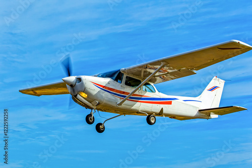 White single engine, high wing aircraft with red and blue stripes flying against a cloudy blue sky