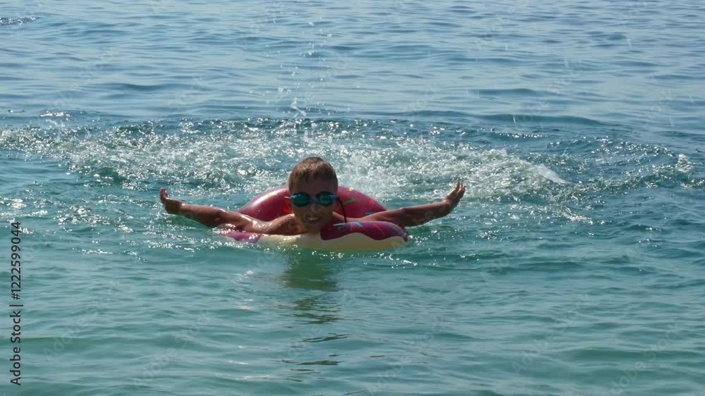 Happy boy floating on colorful swim ring in crystal-clear sea water while wearing goggles on bright summer day. Concept of outdoor fun, carefree childhood, and seaside vacation.High quality 4k footage Happy boy floating on colorful swim ring in crystal-clear sea water while wearing goggles on bright summer day. Concept of outdoor fun, carefree childhood, and seaside vacation.High quality 4k footage