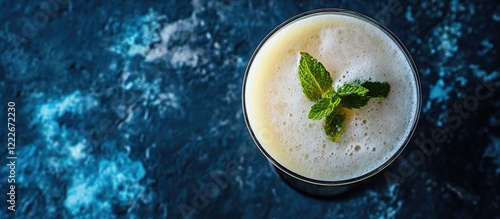 Refreshing Yuzu Kolada cocktail in a clear glass with mint leaves on top against a dark stone surface featuring vibrant blue and black hues. photo