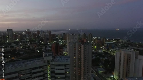 A densely populated coastal cityscape at dusk, with high-rise buildings and glowing city lights extending towards the ocean. The scene reflects urban growth, overpopulation, and environmental impact.