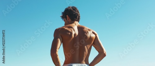 A muscular man stands with his back to the camera against a clear blue sky.