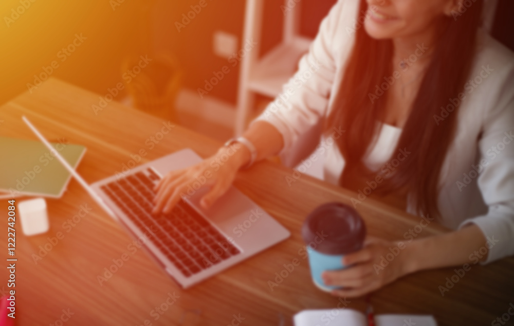 Obraz premium A blurred image of a woman working on a laptop in a cozy home office, holding a coffee cup in one hand, with notebooks and a wireless earphone case on the table, showcasing a productive atmosphere.