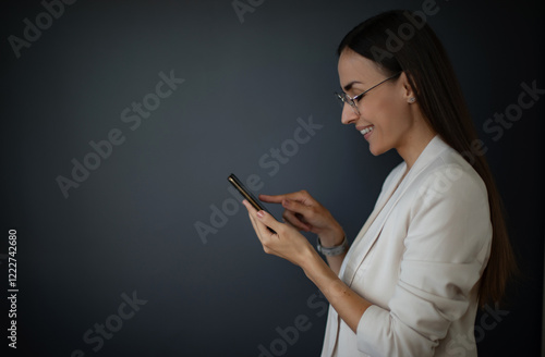 A confident businesswoman with glasses smiles while using her smartphone in a modern office. She's engaged and focused, with a professional attire and a minimalistic, sleek workspace in the background photo