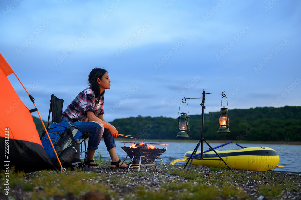 Φωτογραφία Person grilling food by a lakeside, sitting next to an orange tent and camping gear, with lanterns and a yellow kayak adding to the peaceful evening atmosphere