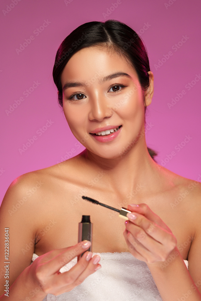 Woman applying mascara while smiling in a beauty setting with soft pink background Woman applying mascara while smiling in a beauty setting with soft pink background