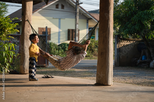 A cheerful boy interacts with his father resting on a hammock under a wooden porch in a rural Thai setting during sunset.