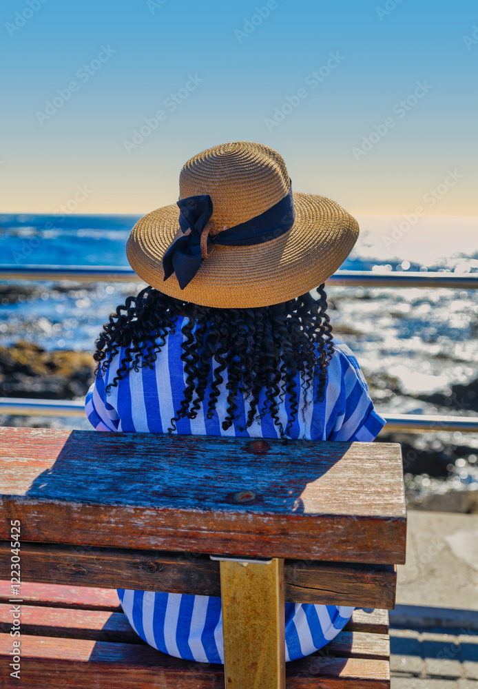African woman in a blue and white striped dress sitting on a bench, enjoying the sea view at Green Point, Cape Town, with the Atlantic Ocean in the background, South Africa