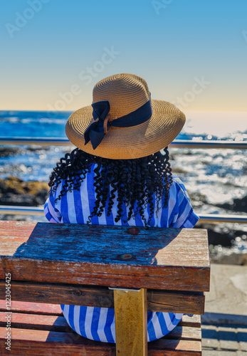 Obraz na płótnie African woman in a blue and white striped dress sitting on a bench, enjoying the sea view at Green Point, Cape Town, with the Atlantic Ocean in the background, South Africa w salonie