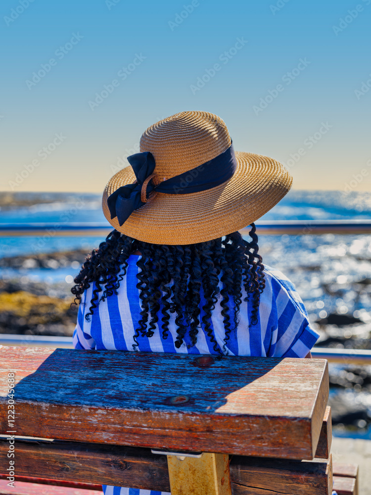 Vertical close-up shot of an African woman in a blue and white striped dress sitting on a bench, enjoying the sea view at Green Point, Cape Town, South Africa