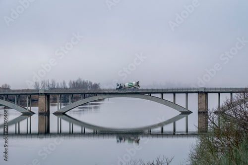 A concrete mixer truck crosses a misty bridge, reflecting on the calm water below as the early morning fog envelops the scene, creating a tranquil and serene atmosphere. photo