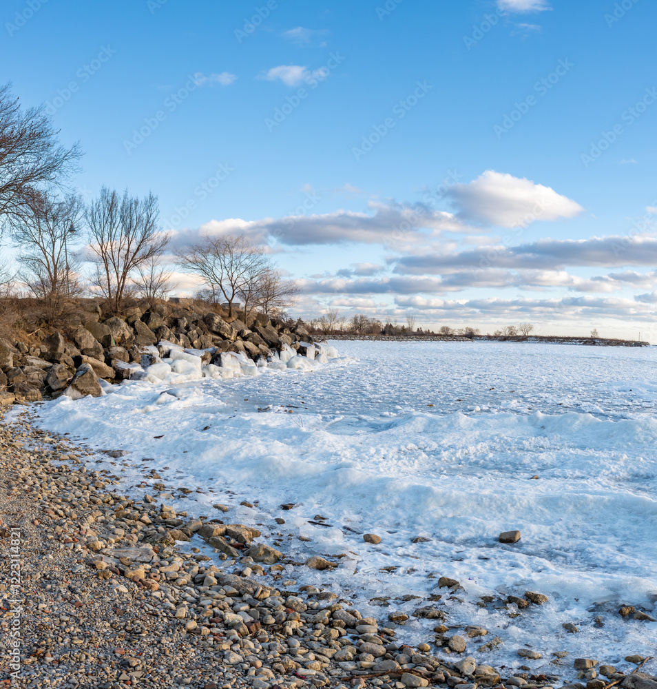 Icy waves crash against a rocky shoreline on a cold winter day. The frozen water contrasts with the clear blue sky, creating a serene yet powerful scene. Bare trees line the shore. Icy waves crash against a rocky shoreline on a cold winter day. The frozen water contrasts with the clear blue sky, creating a serene yet powerful scene. Bare trees line the shore.