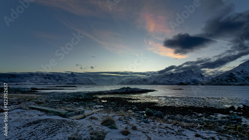 Wide angle view of a fjord surrounded by snow-coverds mountains, and remains of the concrete foundation of a boathouse on rocky shore covered by snow in a cold afternoon. Lofoten, Norway.