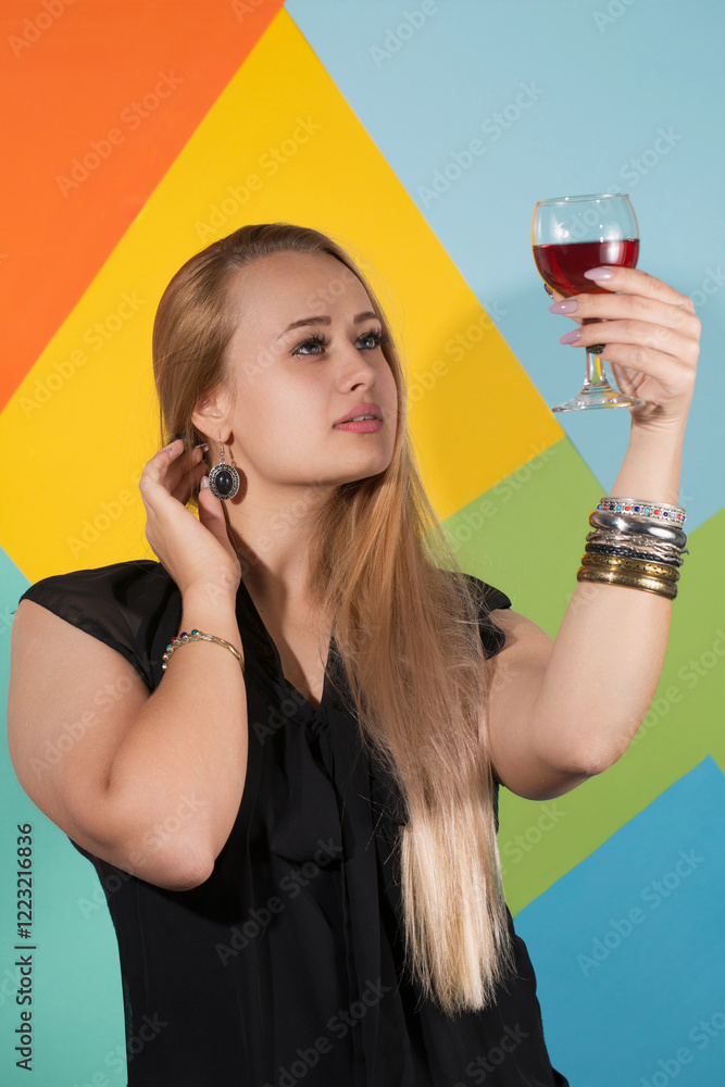 Beautiful blonde in a black dress and metal bracelets holding a glass of red wine and looking at the drink
