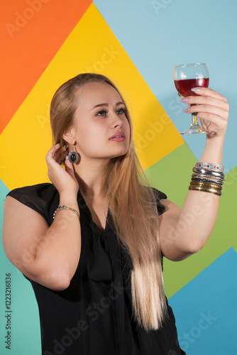 Obraz na płótnie Beautiful blonde in a black dress and metal bracelets holding a glass of red wine and looking at the drink w salonie