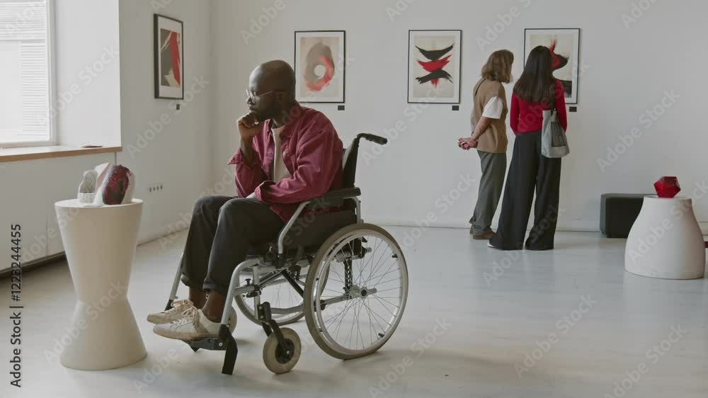 African American man in wheelchair looking thoughtfully at sculpture on pedestal, while other people viewing abstract paintings in wall in minimalist art gallery African American man in wheelchair looking thoughtfully at sculpture on pedestal, while other people viewing abstract paintings in wall in minimalist art gallery