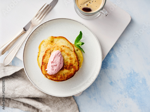 Soft pancakes are plated with a generous dollop of berry cream, garnished with mint, alongside a cup of coffee on a serene light blue countertop photo