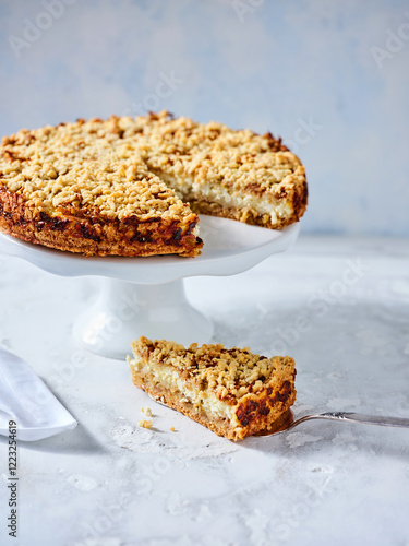 A freshly baked crumbly cake is presented on a white pedestal, showcasing a perfectly cut slice beside it. The setting is bright, inviting, and perfect for dessert lovers photo