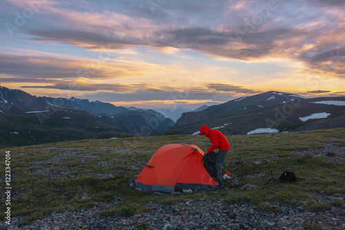 Travel man in vivid red jacket near orange tent on grassy stony pass with view to big mountain range silhouette far away under clouds in colorful sunset color sky. Alpine adventure. Scenic cloudy sky. photo