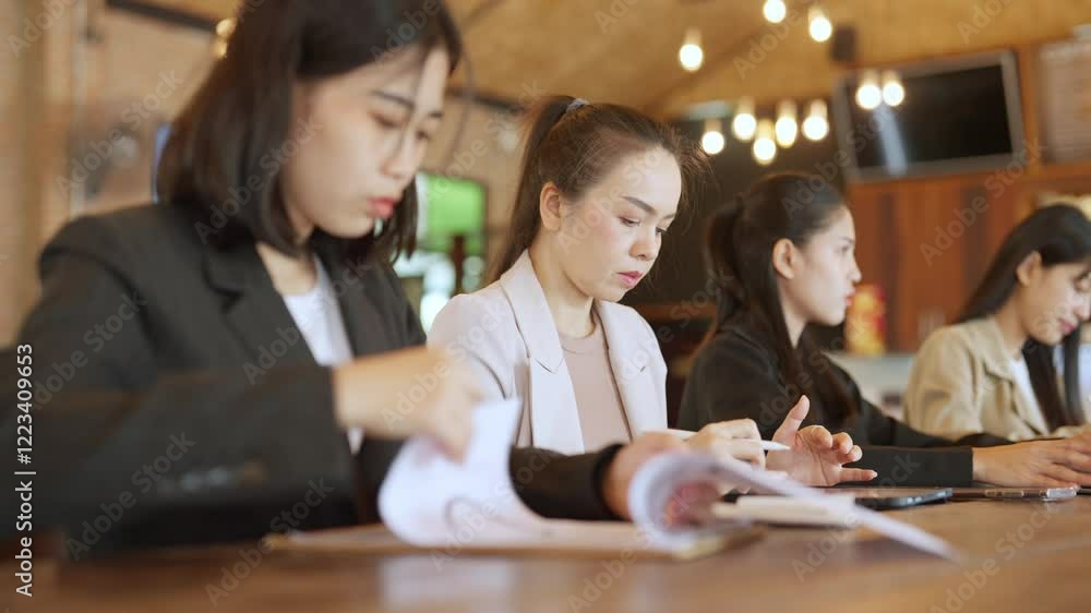 A group of women are sitting at a table, each with a book in front of them. They appear to be focused on their work, possibly studying or working on a project. The atmosphere seems to be serious A group of women are sitting at a table, each with a book in front of them. They appear to be focused on their work, possibly studying or working on a project. The atmosphere seems to be serious