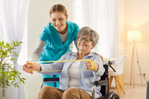 Nurse helping elderly disabled woman with rehabilitation shows how to do physical exercises. Old female pensioner streches arms with resistance band at rehab centre poses in wheelchair  photo