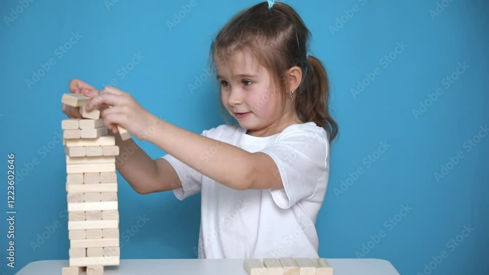 Slow motion footage little girl playing a jenga board game on blue background. Happy child takes wooden blocks from tumble tower and it's falling down. Children entertainment and creativity concept. 