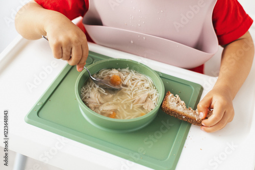 Close-up of a child s hands holding a spoon and a slice of bread while eating chicken noodle soup from a green bowl, illustrating a wholesome and comforting meal photo