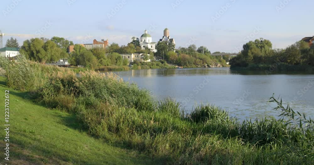A peaceful riverside scene with lush greenery, a bridge, and historic buildings, including a church with a dome. The calm water reflects the serene atmosphere of the sunny day. A peaceful riverside scene with lush greenery, a bridge, and historic buildings, including a church with a dome. The calm water reflects the serene atmosphere of the sunny day.