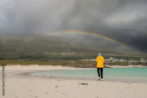 Teenager in yellow jacket walking on a white stone beach by turquoise ocean. Dog bay, county Galway, Ireland. Travel and tourism. Stunning Irish landscape scene. Enjoy nature theme. photo