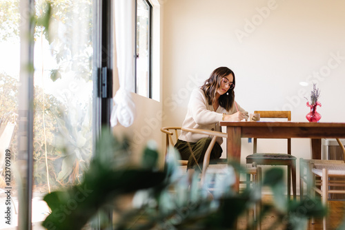 Woman sitting at a table writing in a notebook, surrounded by plants and natural light.