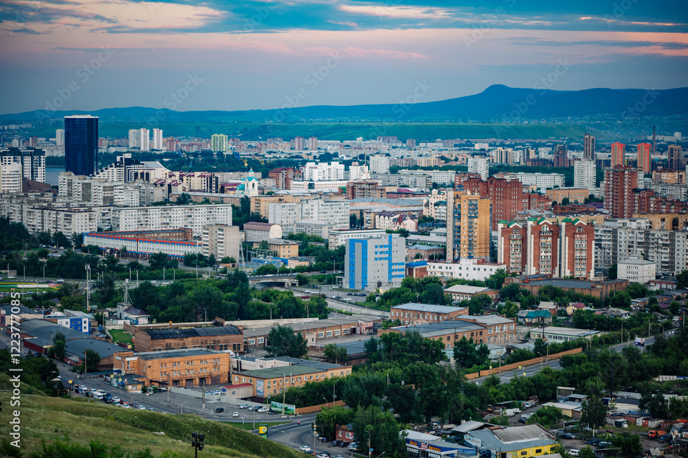 Obraz premium Krasnoyarsk, russia, city skyline featuring residential and industrial buildings at sunset with a mountain in the background