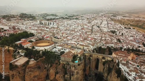 Aerial view capturing Ronda, Spain, featuring the iconic Puente Nuevo bridge, Plaza de Toros bullring, and traditional white houses against a dramatic landscape, drone establishing shot