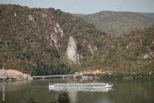 Majestic sculpture of Decebalus carved into cliff greets visitors on Danube River. Cruise ship glides smoothly along tranquil Danube River. Lush green hills adorned with autumn colors frame water photo