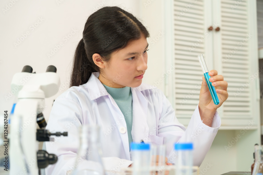 Cute Caucasian schoolgirl wearing white scientist outfit holding test tube filled with blue liquid in science classroom. Curious expression, analyzing chemical reaction, lab equipment and microscope. Cute Caucasian schoolgirl wearing white scientist outfit holding test tube filled with blue liquid in science classroom. Curious expression, analyzing chemical reaction, lab equipment and microscope.