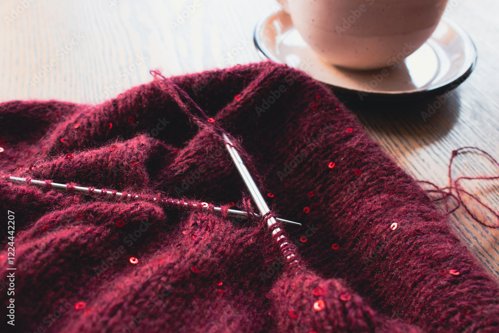 Red knitted sweater with sequins and knitting needles with coffee cup on table. Sequin pattern in needlework with cappuccino. Knitting hobby. Knitted fashion concept. Sequins pattern in knitwear.