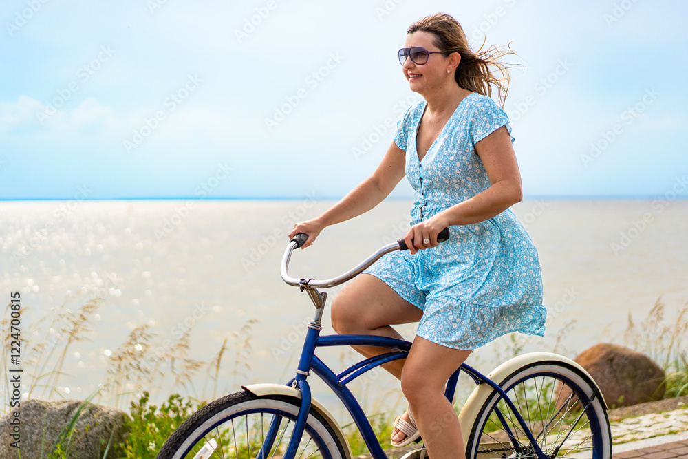 Active summer holidays. Mature woman wearing dress riding blue cruiser bike along watershore in summertime. Side view Active summer holidays. Mature woman wearing dress riding blue cruiser bike along watershore in summertime. Side view