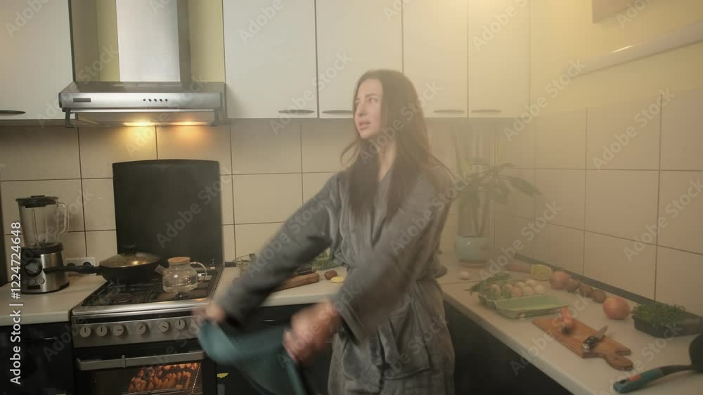 A housewife in a soft gray bathrobe frantically waves a towel to disperse smoke from burnt food in a cozy kitchen filled with various ingredients. A housewife in a soft gray bathrobe frantically waves a towel to disperse smoke from burnt food in a cozy kitchen filled with various ingredients.