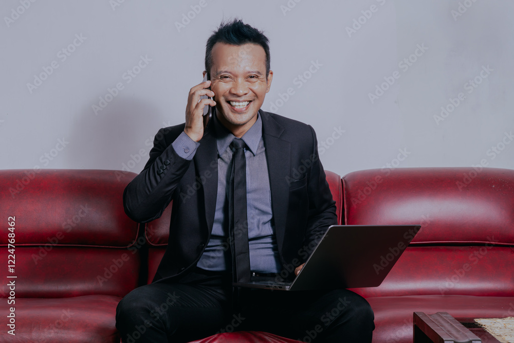 Smiling asian boss manager businessman looking at camera sitting on sofa while talking on mobile phone at the office. Smiling asian boss manager businessman looking at camera sitting on sofa while talking on mobile phone at the office.