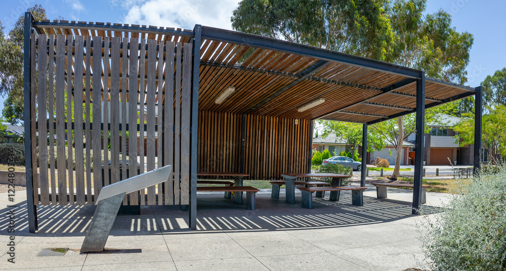 A modern outdoor public shelter or pavilion with a contemporary architectural design and vertical wooden slats. A stylish community rest area with picnic table and built-in seating in a suburban park.