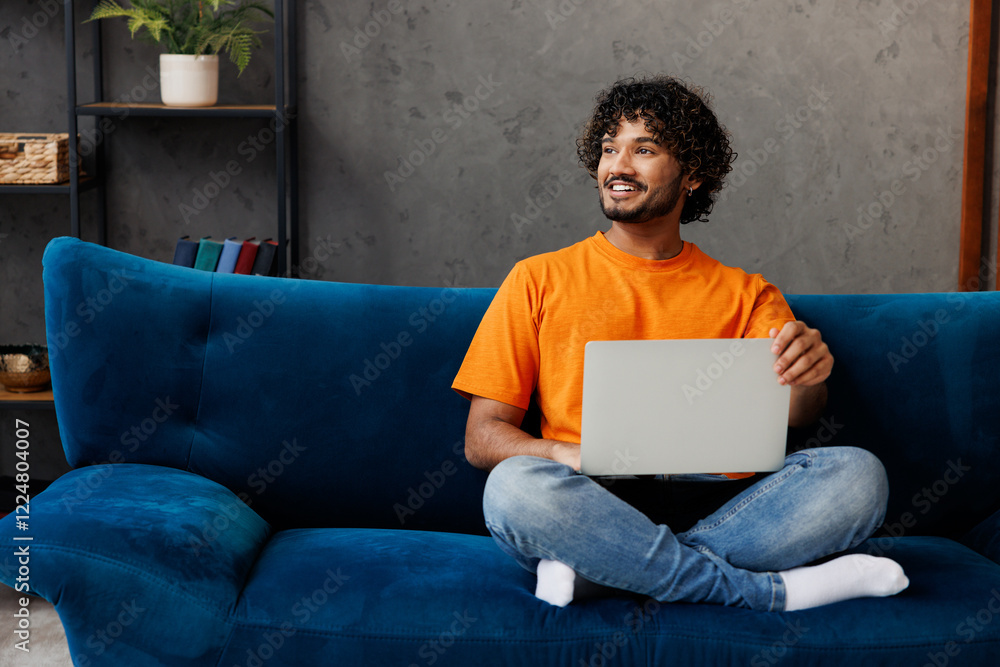 © ViDi Studio - Young IT Indian man wear orange casual clothes sit on blue sofa couch hold use work on laptop pc computer stay home hotel flat rest relax spend free spare time in living room indoor. Lounge concept.