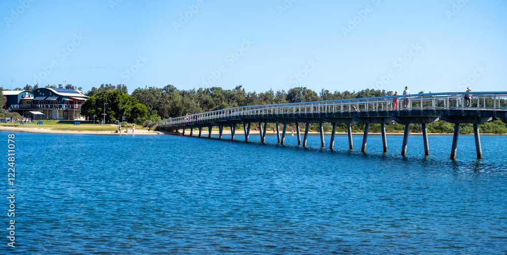 Panoramic view of the Cunninghame Arm Footbridge stretches across the calm blue water of Lake Entrance. People stroll along the bridge, enjoying the scenic views and the summer holiday Panoramic view of the Cunninghame Arm Footbridge stretches across the calm blue water of Lake Entrance. People stroll along the bridge, enjoying the scenic views and the summer holiday
