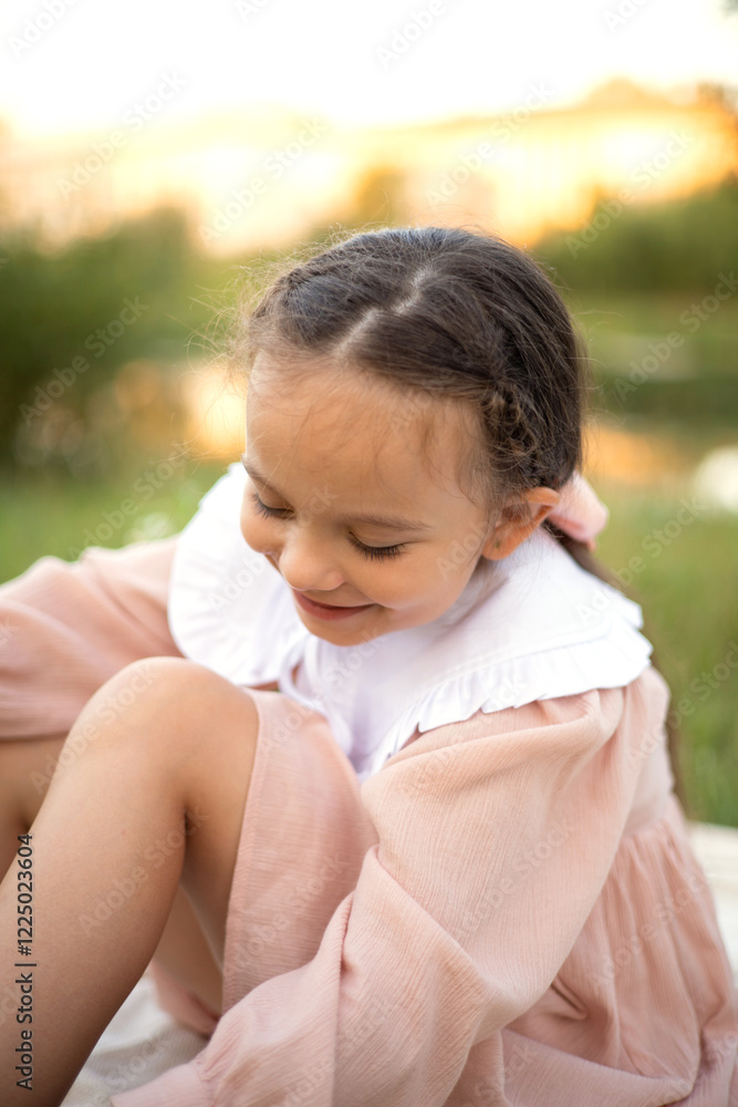 A gentle summer picnic by the water. Portrait of a blue-eyed girl with long hair in a light dress on the grass and on a beige blanket with flower and fruit. A gentle rest. Warm evening