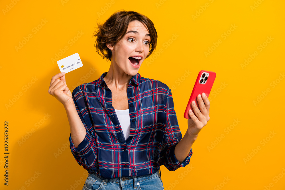 Portrait of excited impressed girl wear plaid shirt staring at smartphone show debit card isolated on bright yellow color background Portrait of excited impressed girl wear plaid shirt staring at smartphone show debit card isolated on bright yellow color background