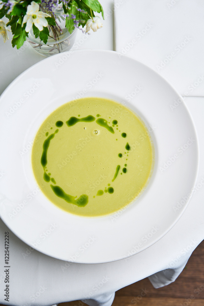 A bowl of creamy green soup is elegantly presented on a white table. Bright drizzles of herb oil create a striking visual on the surface, accompanied by fresh flowers nearby A bowl of creamy green soup is elegantly presented on a white table. Bright drizzles of herb oil create a striking visual on the surface, accompanied by fresh flowers nearby