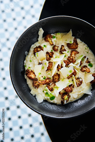 A black bowl holds creamy risotto topped with crispy roasted cauliflower and shavings of cheese. The backdrop features a stylish patterned tablecloth, enhancing the dish's presentation photo