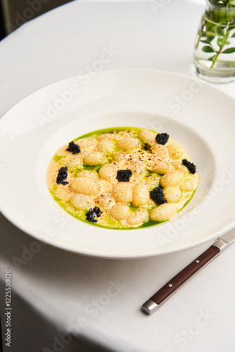 Delicate gnocchi is plated with a vibrant green sauce and topped with caviar, set on a white tablecloth in an upscale dining environment