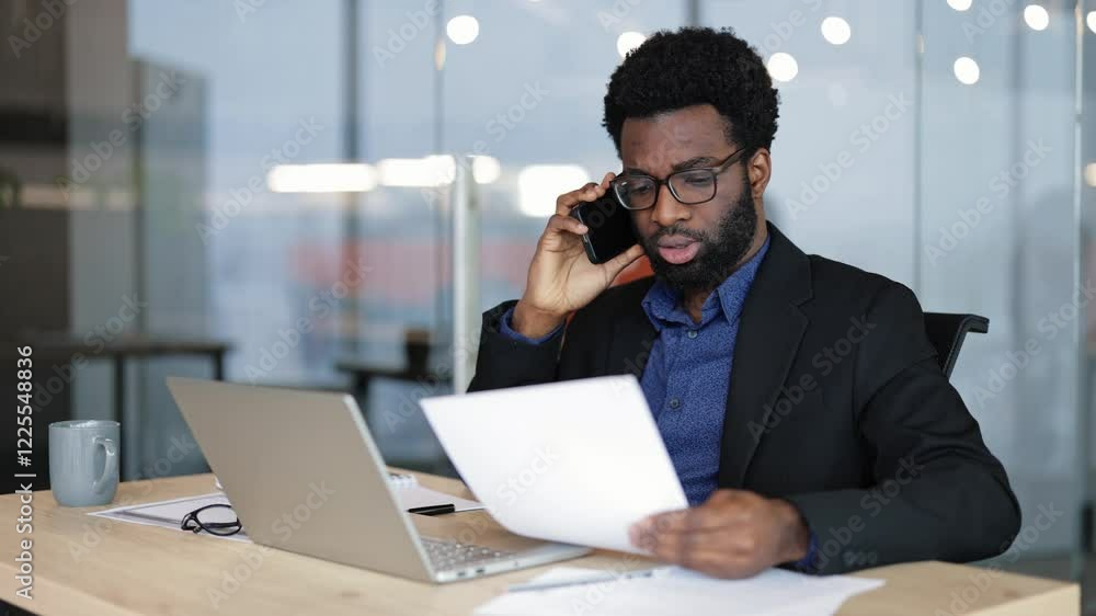 Serious businessman talking on the phone and looking at documents while sitting at his desk. He is working inside a bright office during the daytime. Serious businessman talking on the phone and looking at documents while sitting at his desk. He is working inside a bright office during the daytime.