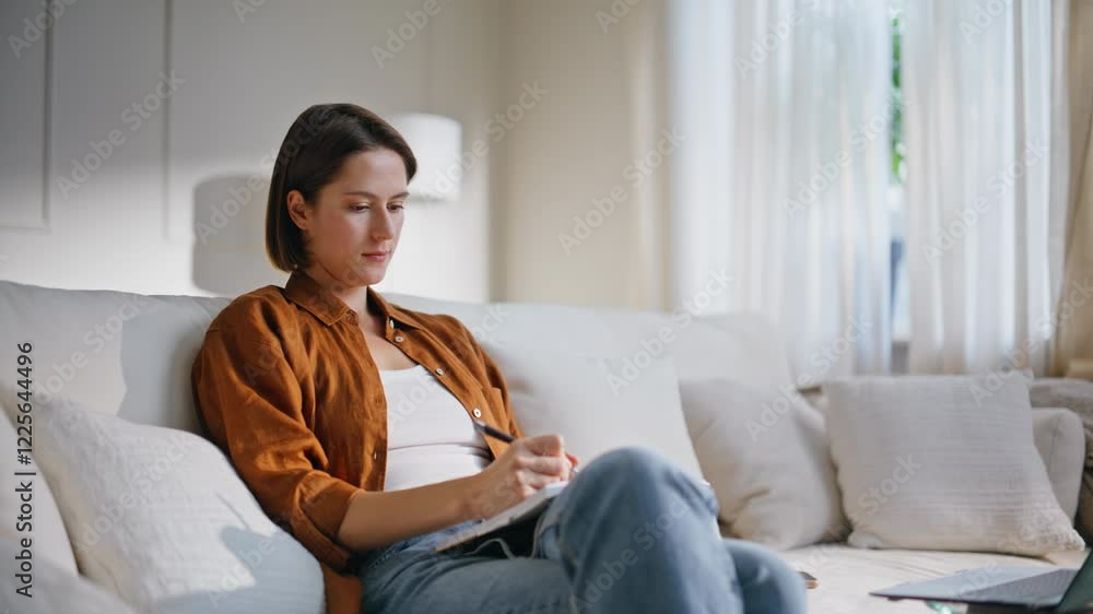 Focused woman writing notepad sitting sofa. Brunette watching webinar at laptop Focused woman writing notepad sitting sofa. Brunette watching webinar at laptop